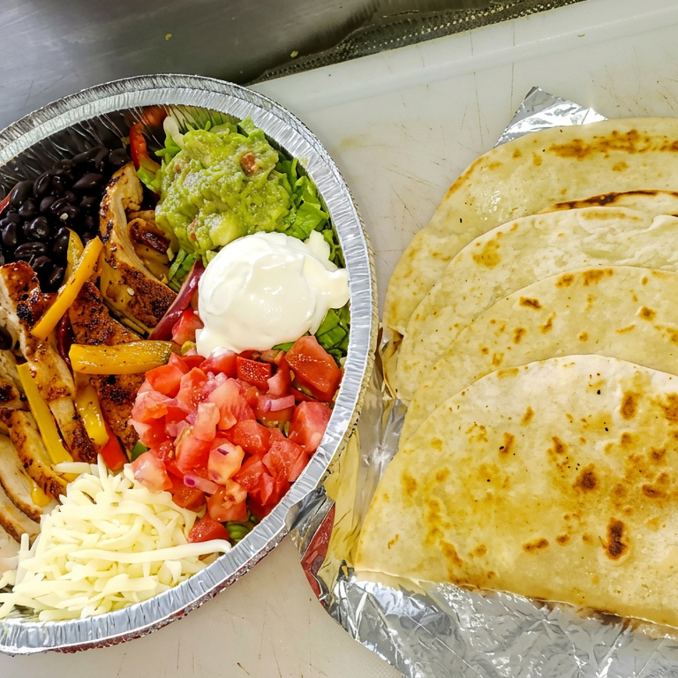 Aluminum bowl of grilled chicken, rice, beans, peppers, guacamole, sour cream, salsa, and cheese beside folded flour tortillas.